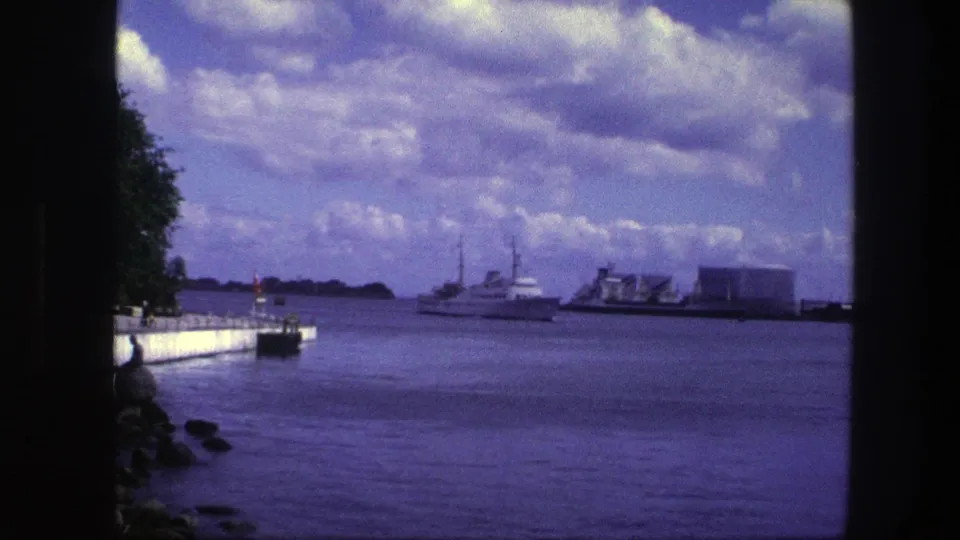 Copenhagen 8mm home-movie harbor view with a passenger boat, industrial waterfront, quay, rocky shore, trees, and blue-purple film color.