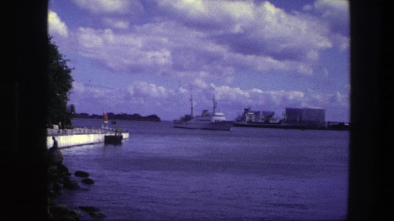 1975 Copenhagen harbor view with a white motorboat crossing calm water, a concrete pier at left, and low industrial buildings in the distance.