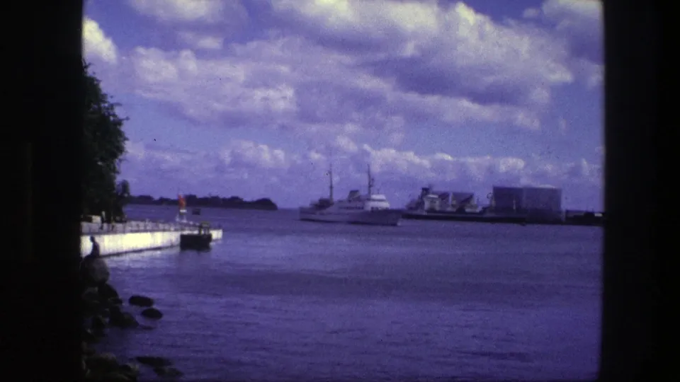 1975 Copenhagen harbor view with a white motorboat crossing calm water, a concrete pier at left, and low industrial buildings in the distance.