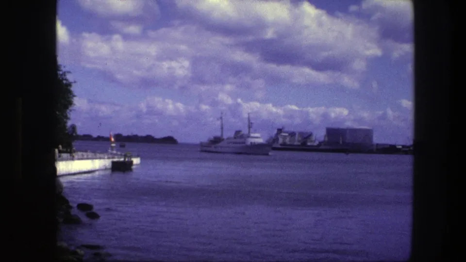 Copenhagen, 1975, with a motorboat passing a distant industrial shoreline under a cloud-filled sky, framed by a dark vertical edge.