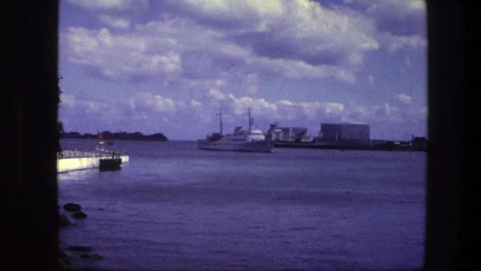 A 1975 Copenhagen harbor frame shows the boat near mid-distance, with rocks and a pier at left and a broad cloudy sky above.