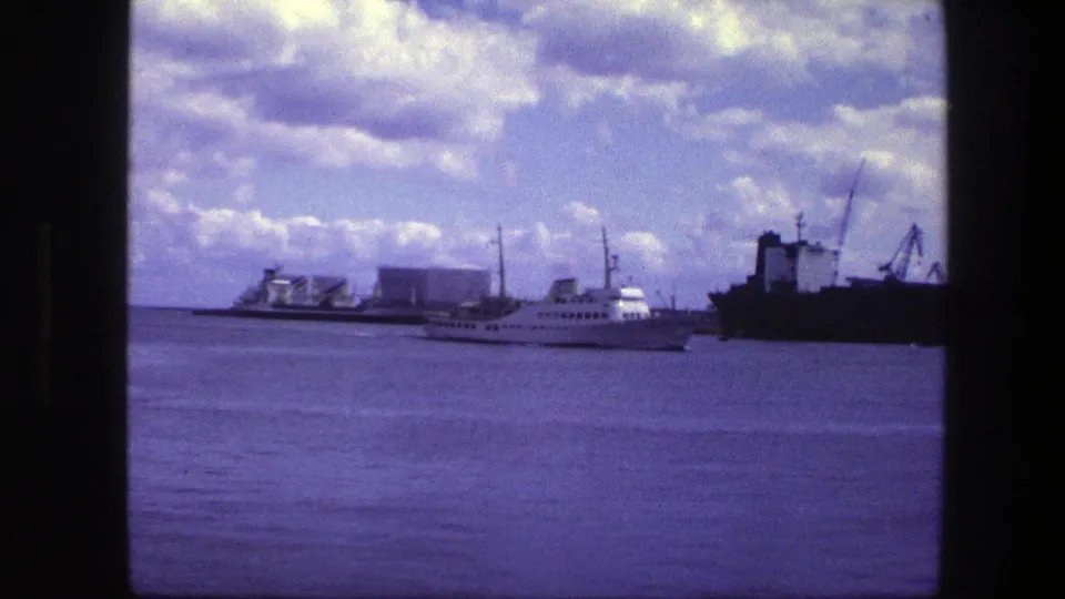 1975 Copenhagen harbor view with a white passenger ship moving past port buildings, cranes, and a dark cargo vessel under cloudy sky.