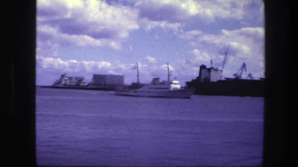 White passenger ship seen at distance in Copenhagen harbor, framed by cloudy sky, port structures, cranes, and a large dark ship.