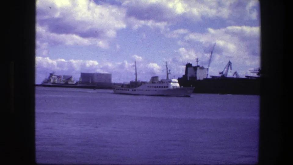 Copenhagen harbor in 1975 with a white passenger vessel crossing in front of a dark cargo ship, cranes, and low waterfront structures.