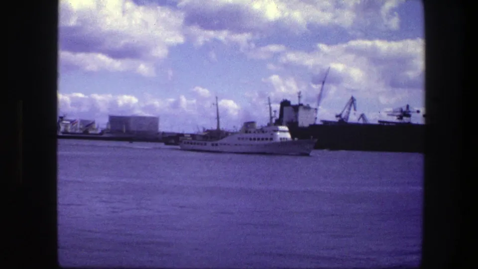 Distant Copenhagen harbor view showing a white passenger ship alongside industrial docks, cranes, and a dark cargo vessel on aged 8mm film.