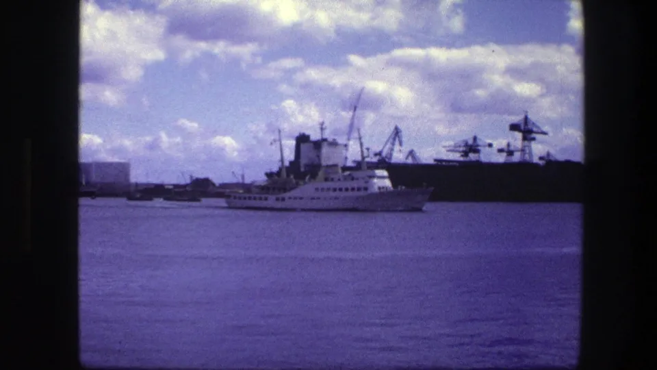 A white cruise ship sits near center frame in 1975 Copenhagen port footage, with cranes and low industrial buildings beyond the water.