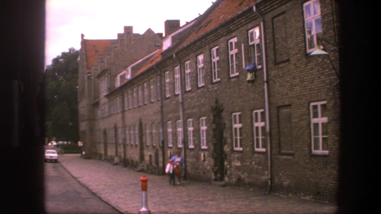 Person with bicycle stands near a brick building as a white car appears down the street.