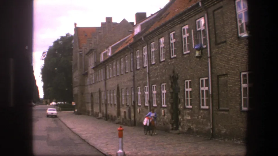 1975 Copenhagen 8mm frame showing a white car on the street and a lone bicycle walker beside the brick building.