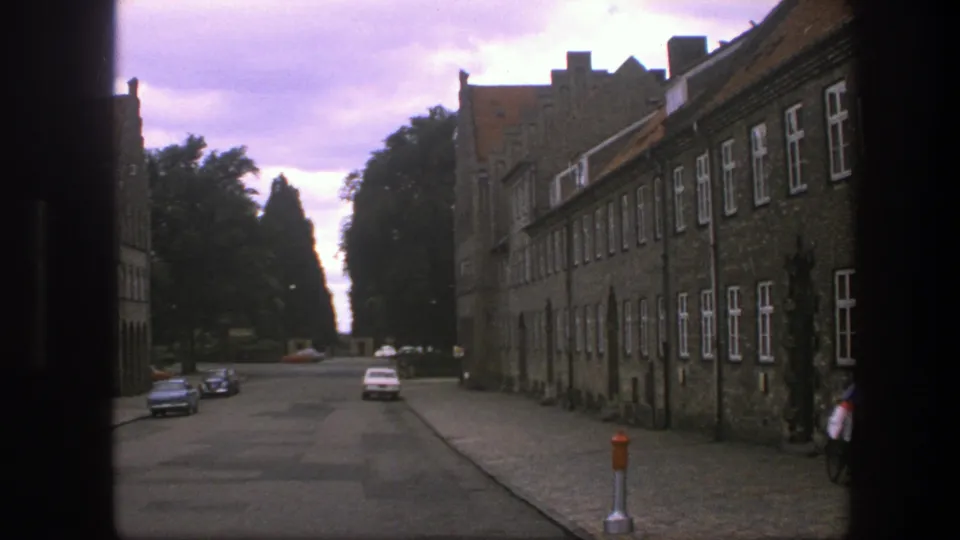 1975 Copenhagen street scene with a long brick building, cobblestone surface, parked cars, trees, and a cyclist near the right edge.