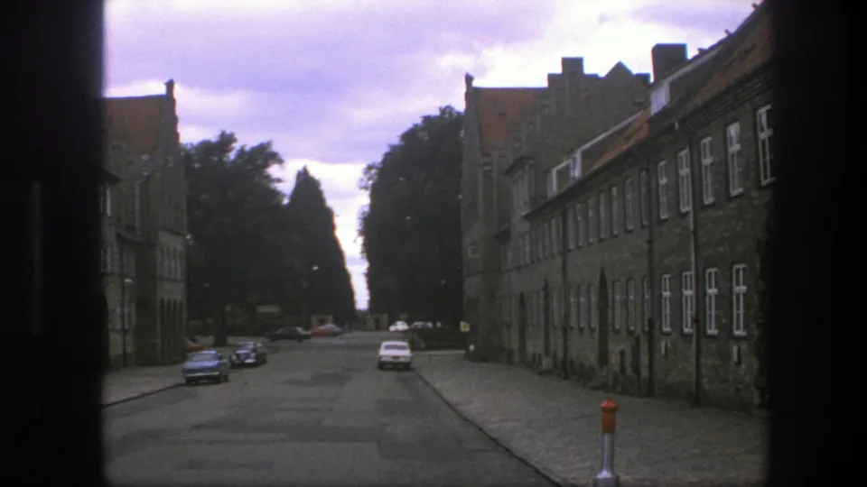 Copenhagen street footage from 1975 showing brick buildings, parked cars, trees, cloudy sky, and the dark edge of the camera framing.