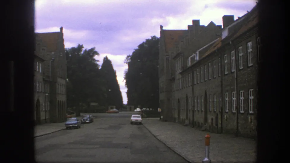 1975 Copenhagen street seen through a dark frame, with stone buildings, cobblestones, parked cars, and trees beyond.