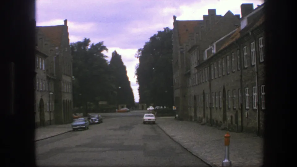 1975 Copenhagen home-movie frame showing a cobbled street, stone facades, parked cars, and dark doorway or window edges.