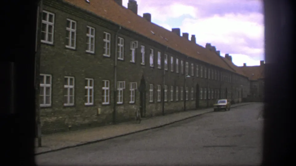 1975 Copenhagen view down a narrow street bordered by a long brick building, white windows, and a parked car.