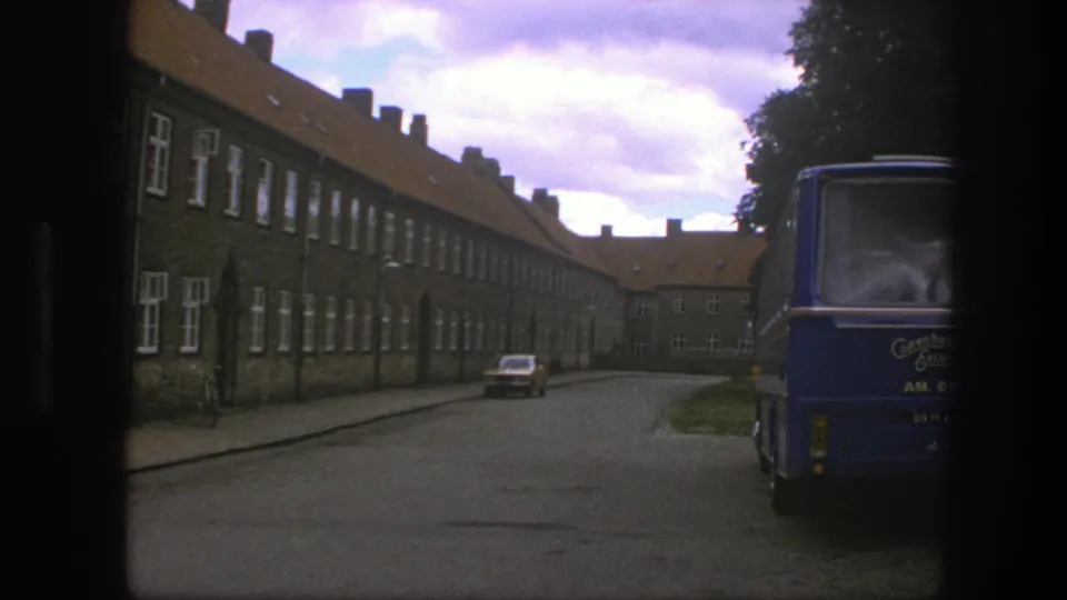 1975 Copenhagen street view with a blue Copenhagen Excursions bus at right, brick buildings, and a small parked car down the road.