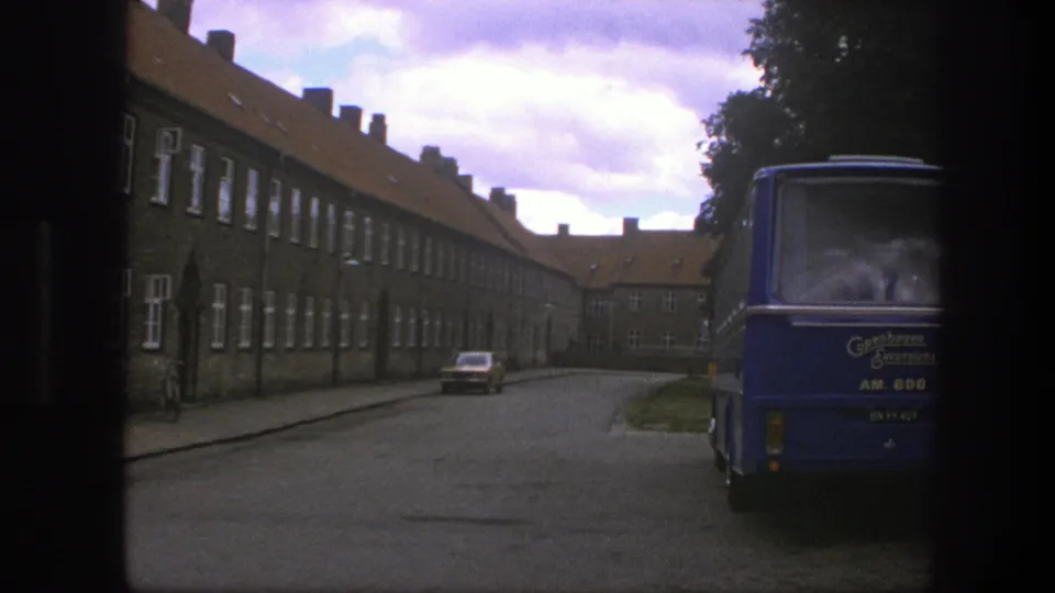 Copenhagen 1975 exterior with a blue excursion bus in the foreground, long brick housing blocks, and a parked car on the quiet road.