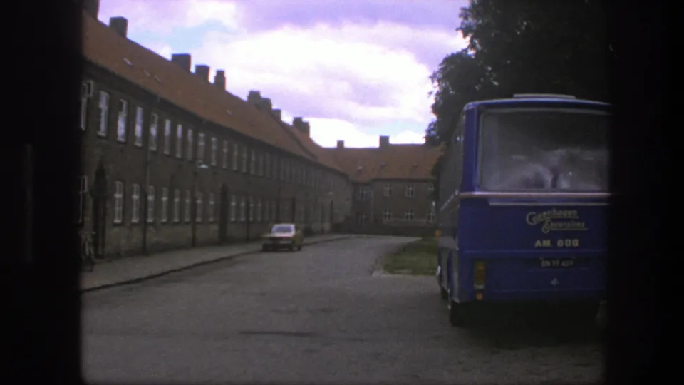 1975 Copenhagen street view with a blue Gentofte-marked bus, red-roofed brick buildings, a parked car, and dark window-frame edges.