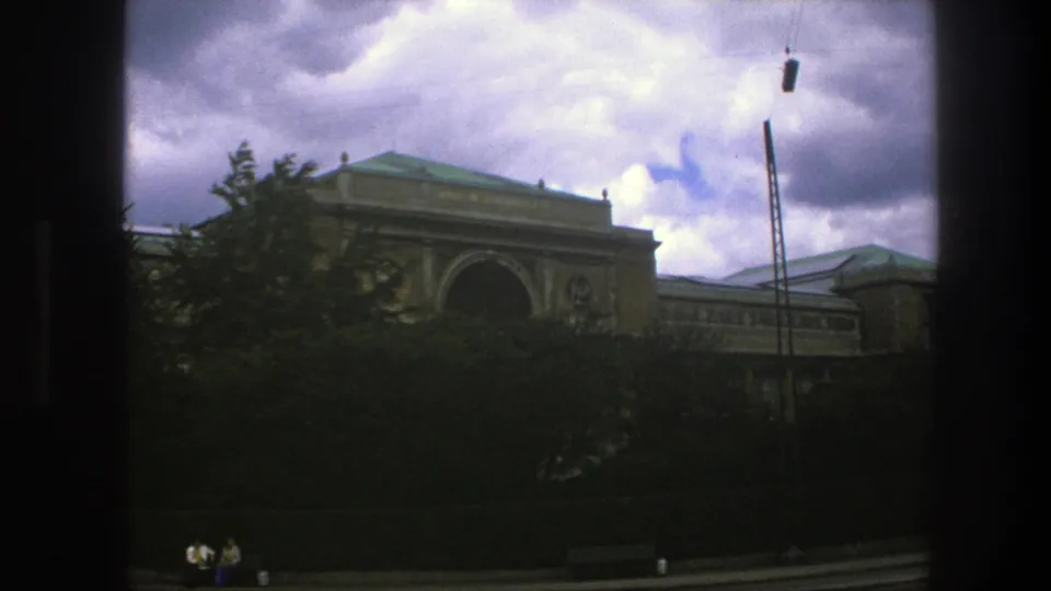 1975 Copenhagen view through a window frame toward a classical-style building with a green roof, arched entrance, trees, and cloudy sky.