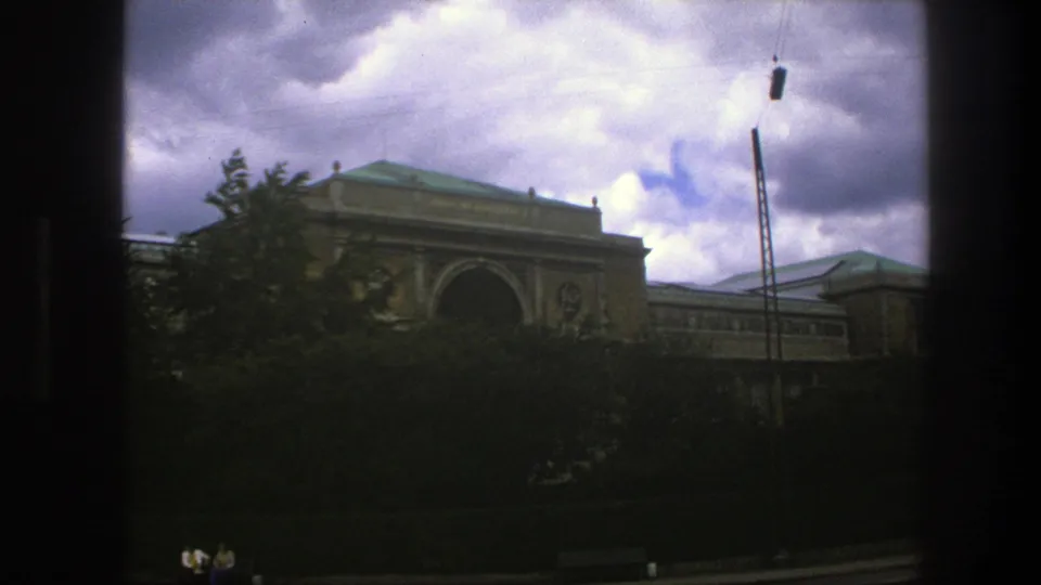 1975 Copenhagen exterior view of a classical building and green roof behind dense trees, with a tall pole and small distant people near the lower edge.
