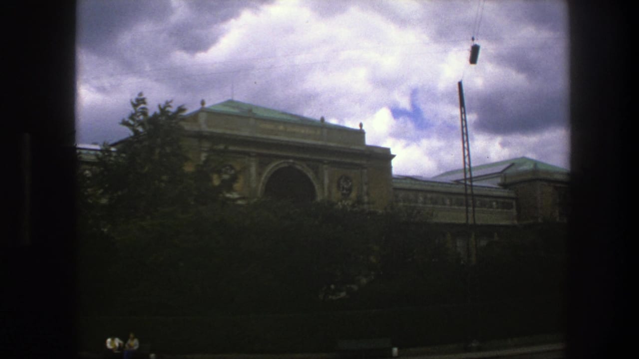 Large tan building partly hidden by trees under a cloudy sky.