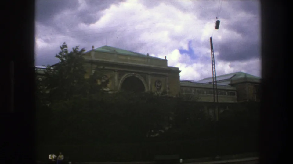 1975 Copenhagen 8mm frame showing a large tan building behind trees, with a tall pole and cloudy sky.