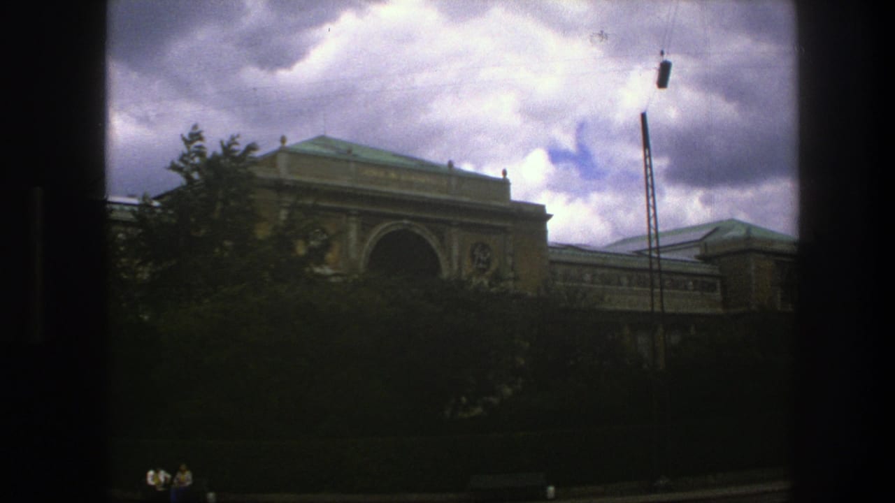 Tan building facade with trees in front and a tall pole against clouds.