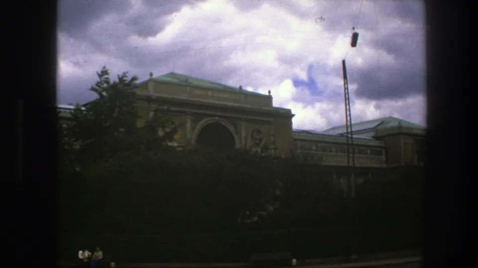 Copenhagen home-movie frame of a tan building and trees, with cloudy sky and visible 8mm frame edges.