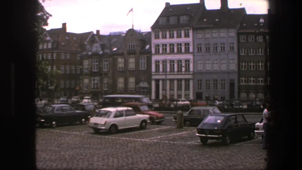 1975 Copenhagen street view with parked cars, cobblestones, gabled buildings, and a Danish flag visible above the roofline.