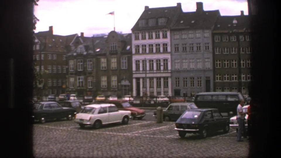 Copenhagen street frame with 1970s-era parked cars, cobblestones, pedestrians, gabled facades, and a Danish flag on a building.