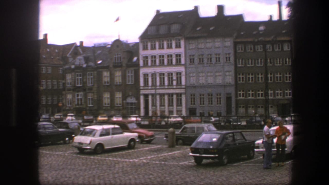 Cobblestone parking lot with 1970s cars, pedestrians, and a row of multi-story buildings.