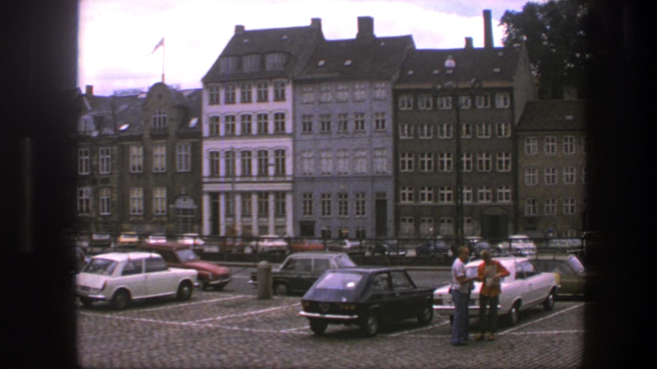Parked cars and pedestrians in a cobblestone lot with historic buildings in the background.