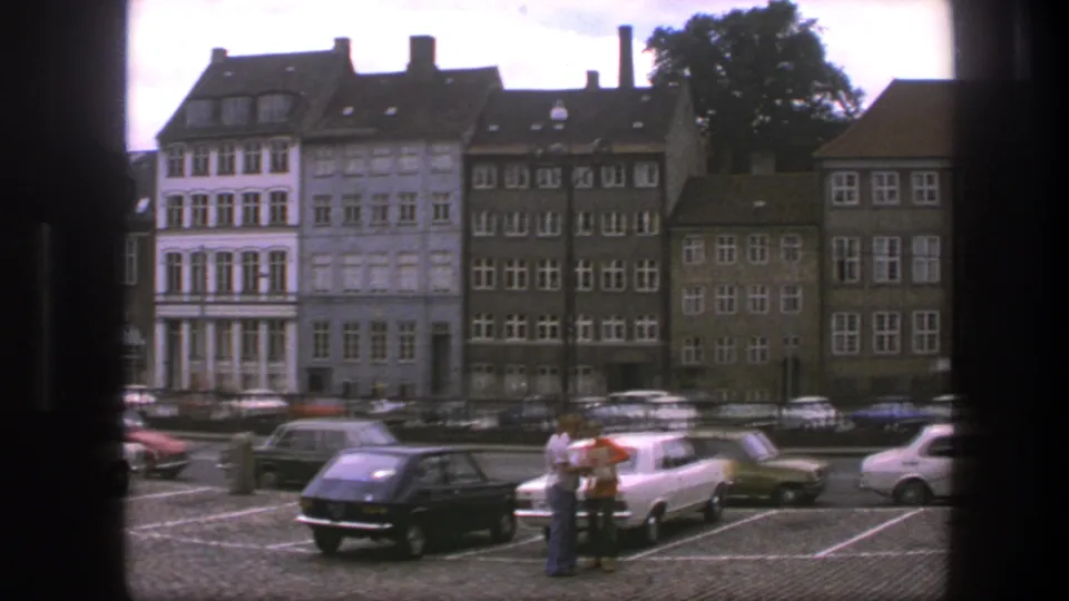 Copenhagen, Denmark, 1975 frame with people near parked compact cars, cobblestones, and older urban buildings across the street.
