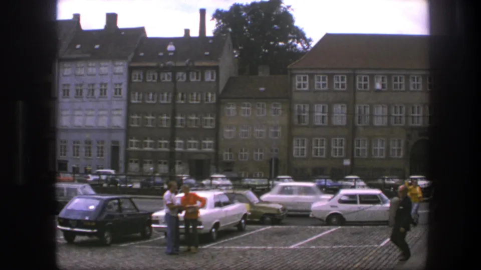 1975 Copenhagen home-movie frame showing a cobblestone parking area, older buildings, several parked cars, and a passerby at right.