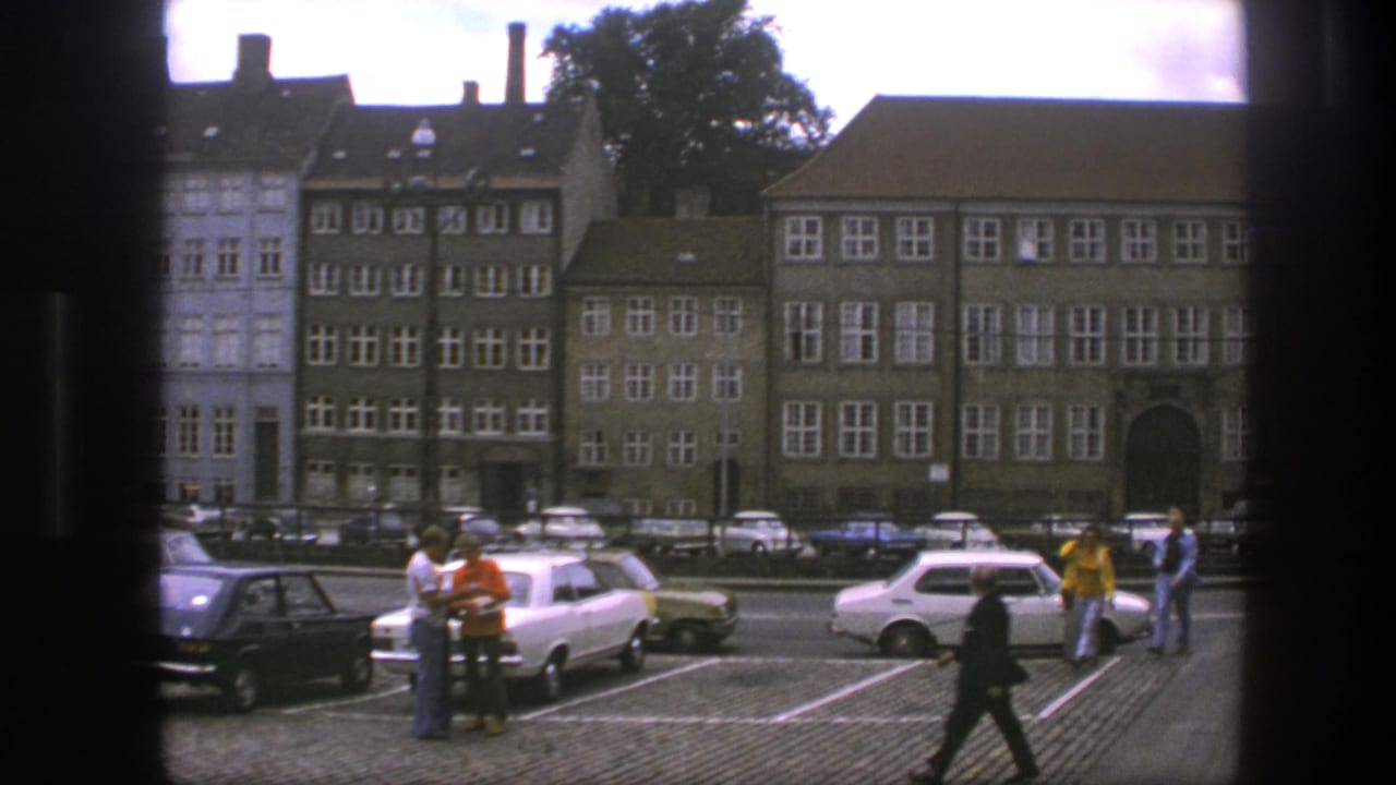 Cobblestone parking area with pedestrians, parked cars, and multi-story buildings across the street.