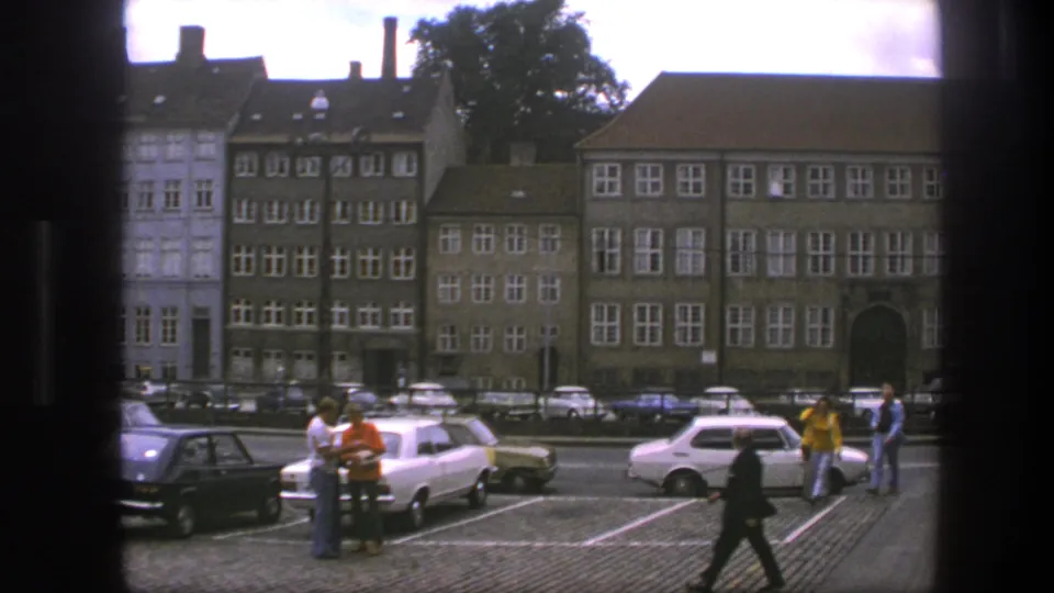 1975 Copenhagen street view with pedestrians near parked cars and large buildings with white-framed windows beyond a cobblestone lot.