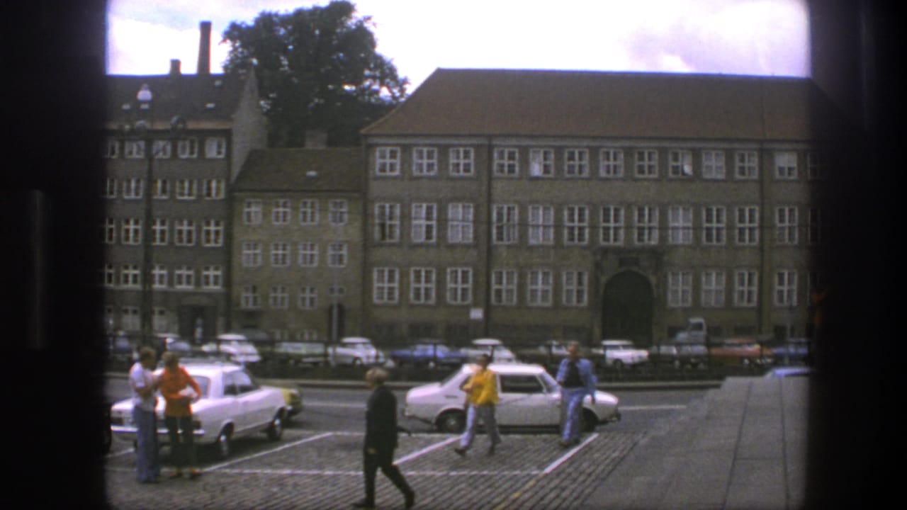 Pedestrians walk through a parking area with compact cars and large buildings in the background.