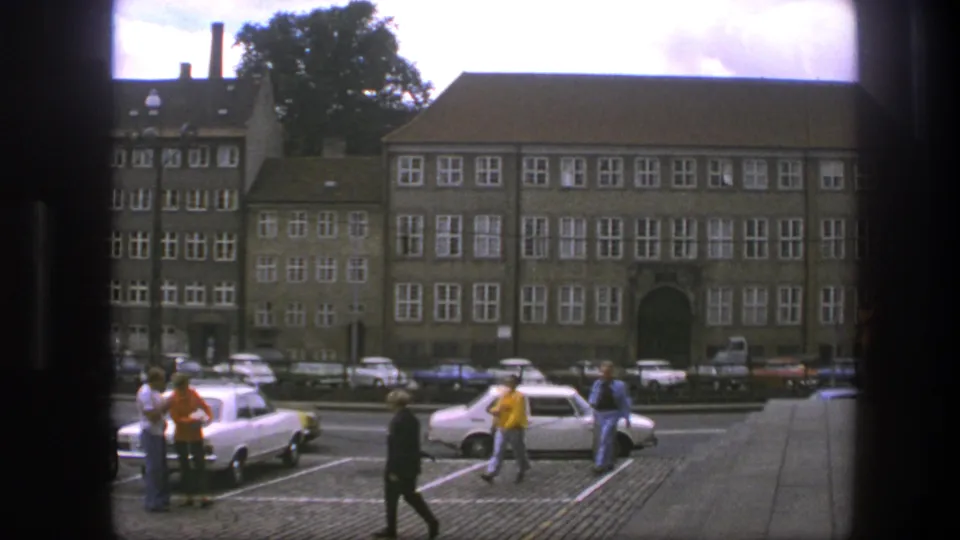 Copenhagen street scene from 1975 showing people in casual clothing moving past parked compact cars and broad institutional buildings.