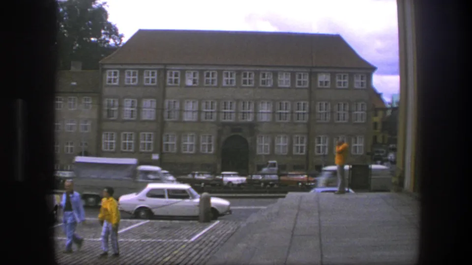 From steps in Copenhagen, the 1975 home movie frames pedestrians below, a white compact car, and a large windowed building across the street.