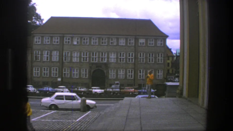 1975 Copenhagen view from broad steps, with a person in a yellow jacket, parked cars, and a large building with many white-framed windows.