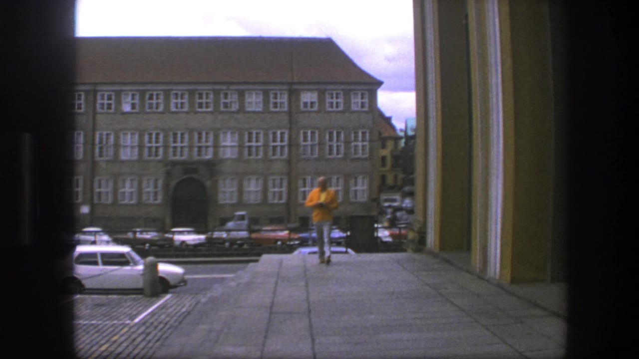 Doorway-framed 1975 Copenhagen street scene with 1970s cars, a broad sidewalk, and a man in a yellow jacket moving away from camera.