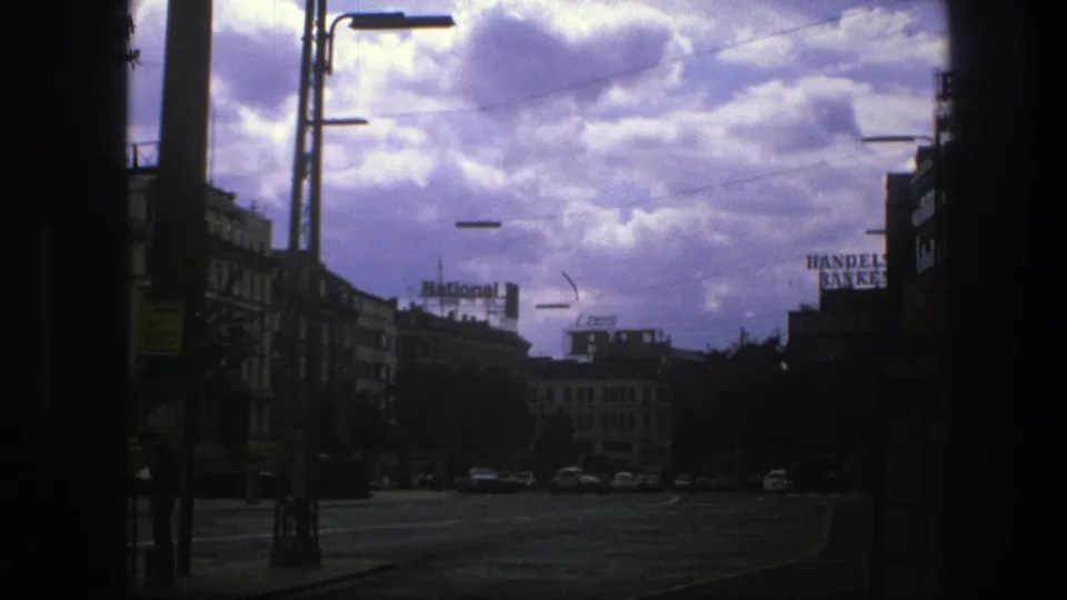 A 1975 Copenhagen street scene shows a broad roadway, older cars, rooftop signs, commercial buildings, and heavy clouds overhead.
