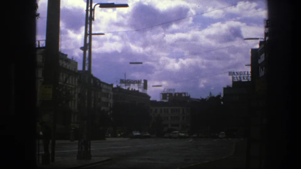 1975 Copenhagen street view under heavy clouds, with dark foreground framing, street lamps, buildings, parked cars, and visible rooftop signs.