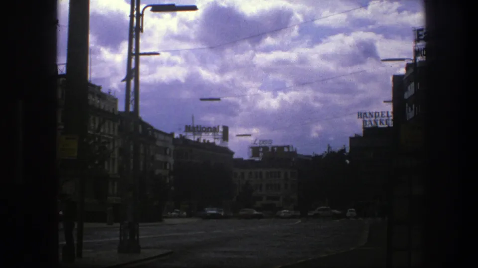 1975 Copenhagen urban street scene with strong cloud cover, silhouetted framing, overhead lamps, distant cars, and readable commercial rooftop signs.