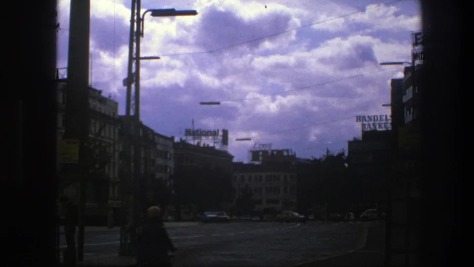 1975 Copenhagen street view with visible commercial signs, parked cars, and a cyclist passing through the lower foreground under a cloudy sky.