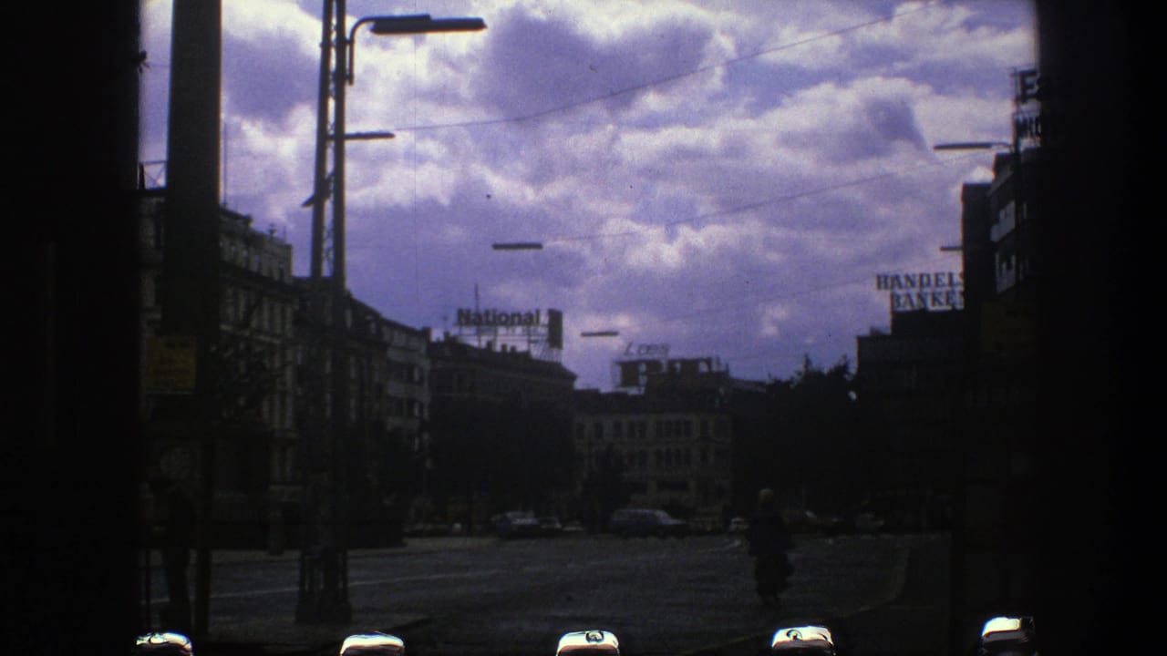 Dark-framed city street with buildings, streetlights, overhead wires, cars, and a cloudy sky.