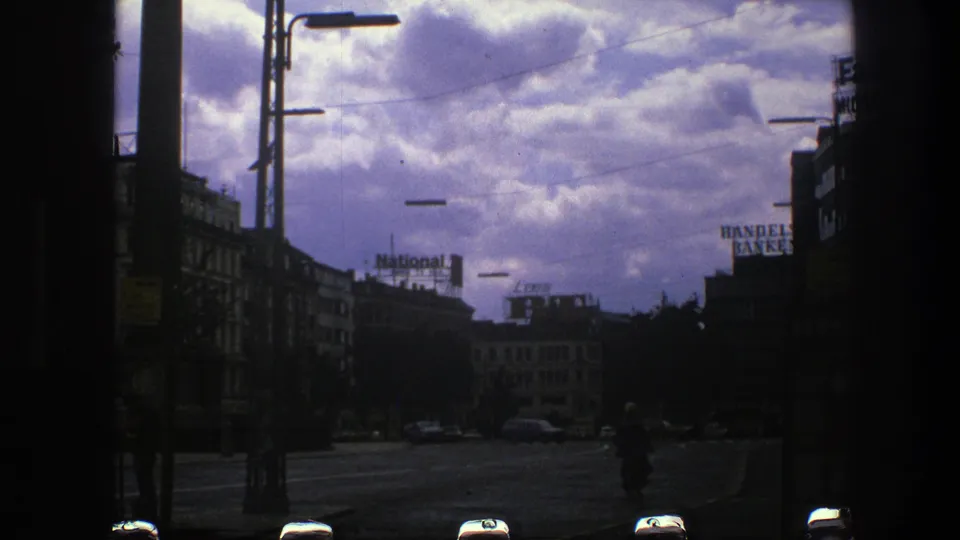 1975 Copenhagen street view on 8mm film, with silhouetted buildings, streetlights, overhead wires, cars, and a dramatic cloudy sky.