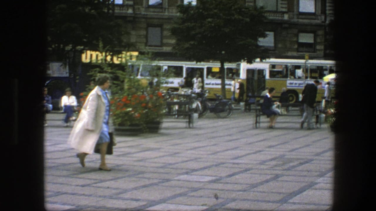 In a Copenhagen plaza, a woman in a light coat and blue dress walks past benches, bicycles, pedestrians, and yellow-and-white buses.