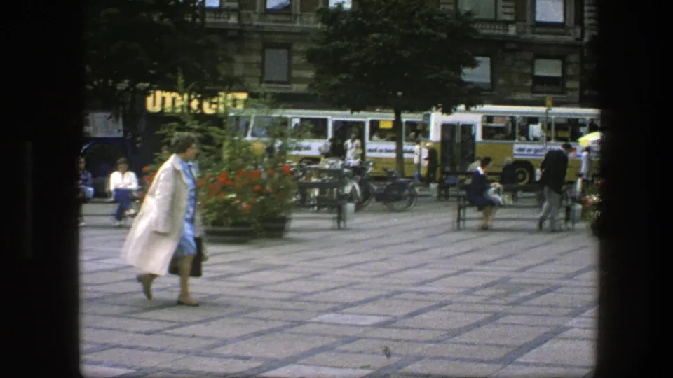 In a Copenhagen plaza, a woman in a light coat and blue dress walks past benches, bicycles, pedestrians, and yellow-and-white buses.