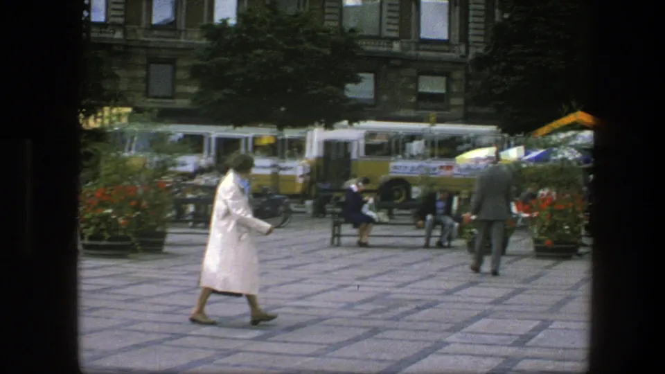 1975 Copenhagen plaza scene with pedestrians crossing tiled paving as buses, benches, bicycles, flowers, and cafe umbrellas appear behind them.