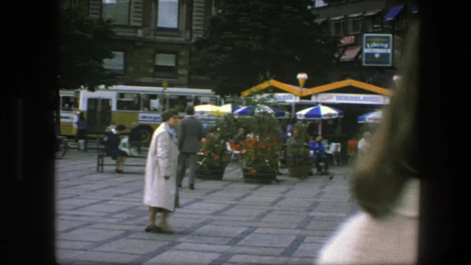 Copenhagen public square with a woman in a light coat near yellow-and-white buses, flower planters, umbrellas, benches, and seated pedestrians.