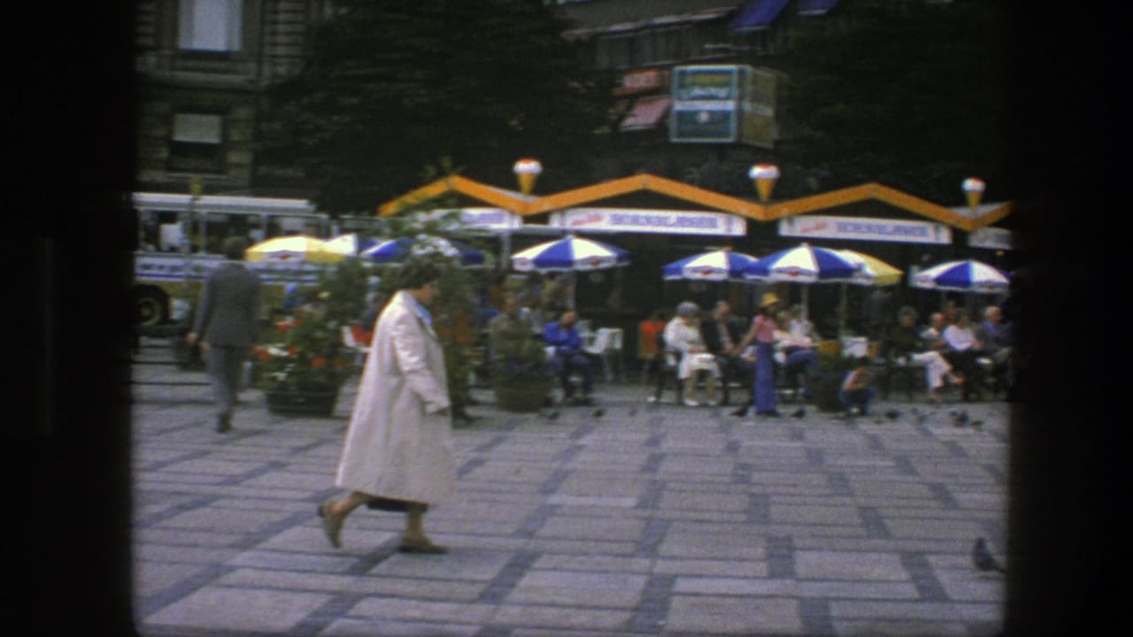 Woman in a white coat walks across a paved square past outdoor cafes and umbrellas.